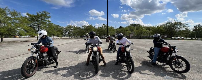 A class of 6 people sitting on motorcycles all facing the same direction and in two lines,  in a parking lot, with all students practicing looking to the left. 