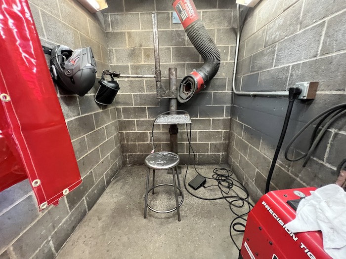 A concrete cinderblock welding booth with a stool, steel table, foot pedal, TIG welding torch and motorcycle helmet hanging on the wall. 