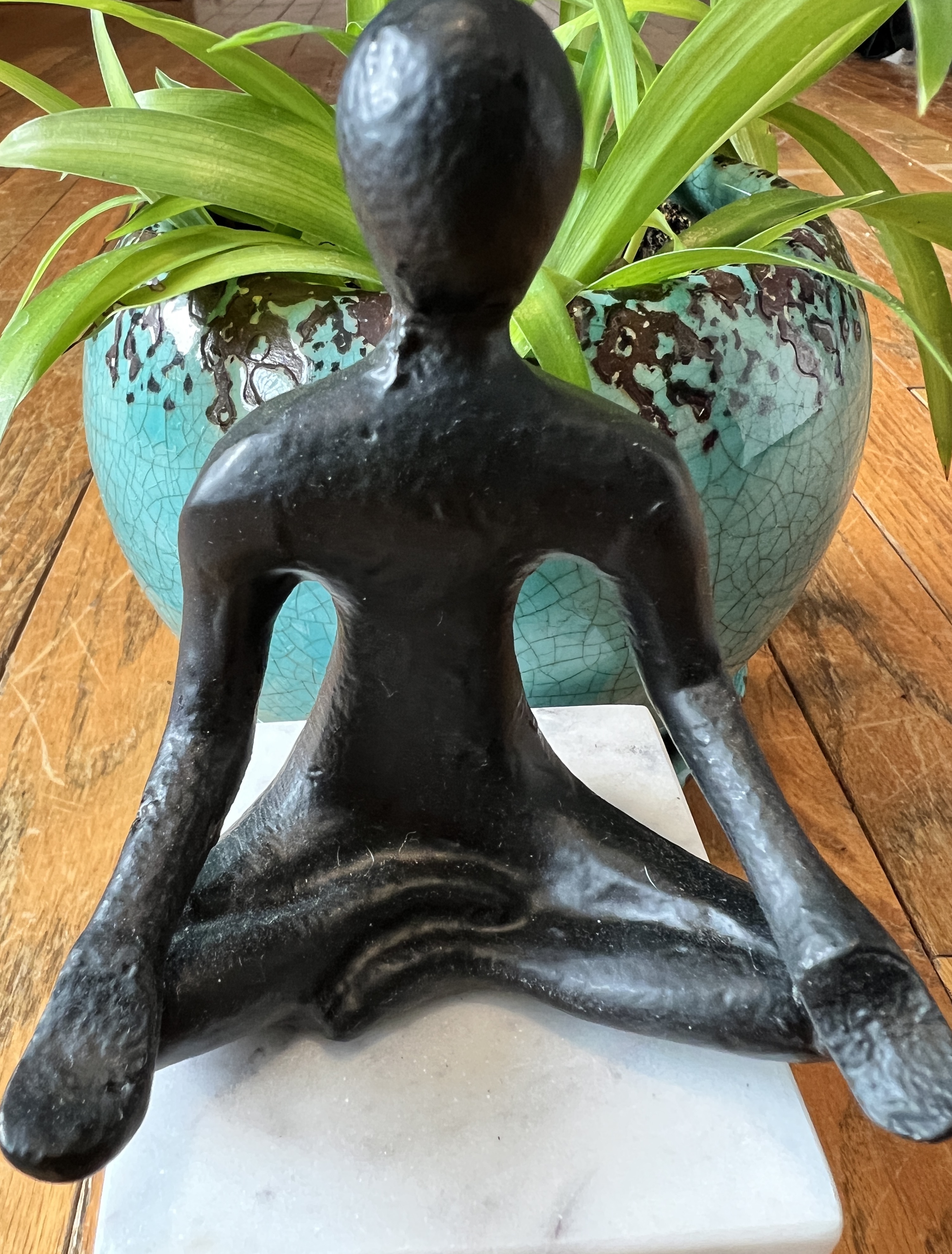 sculpture of a human figure seated cross-legged with hands on knees and a spider plant in a blue pot behind