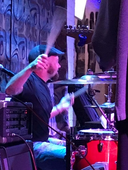 Daniel sitting at a red drumset with his right hand about to hit the ride cymbal and his right hand hitting the snare drum, looking off into the crowd