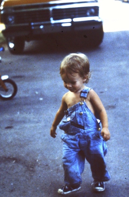 old photo of Daniel as a 2 or 3 year old, walking in denim overalls with one strap falling off, a tricycle front wheel and a 1980's pickup truck in the background. 