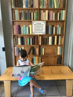 My daughter sitting on a bench with legs crossed, holding a book in front of a wooden wall that looks like a bookshelf with books on it.