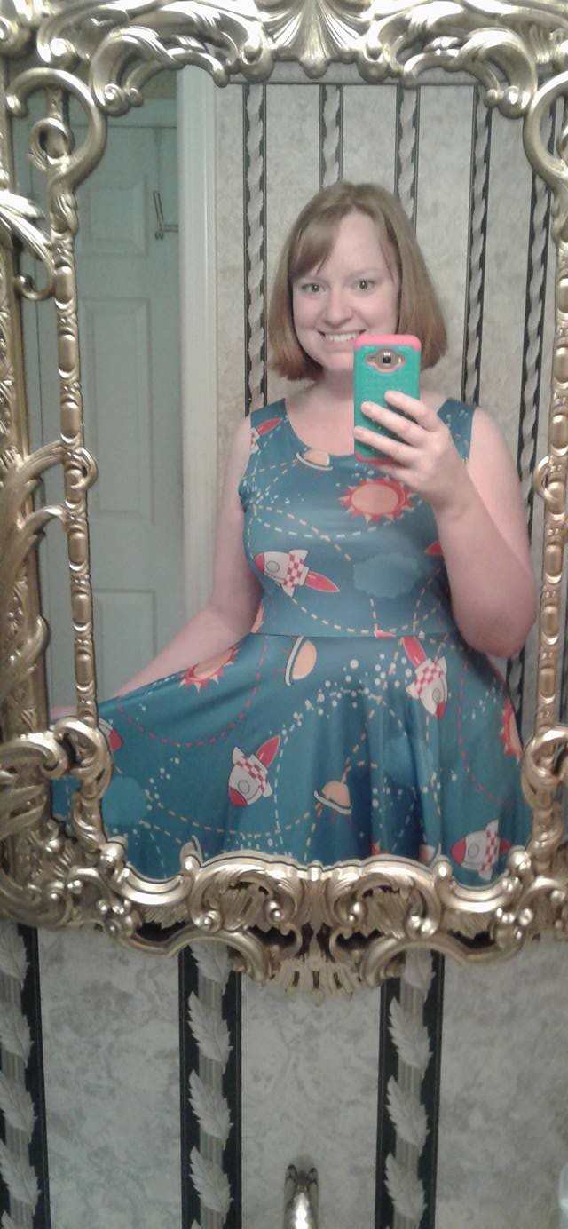 Jessica holding her cellphone taking a selfie in a mirror wearing a blue dress with cartoon rocketships